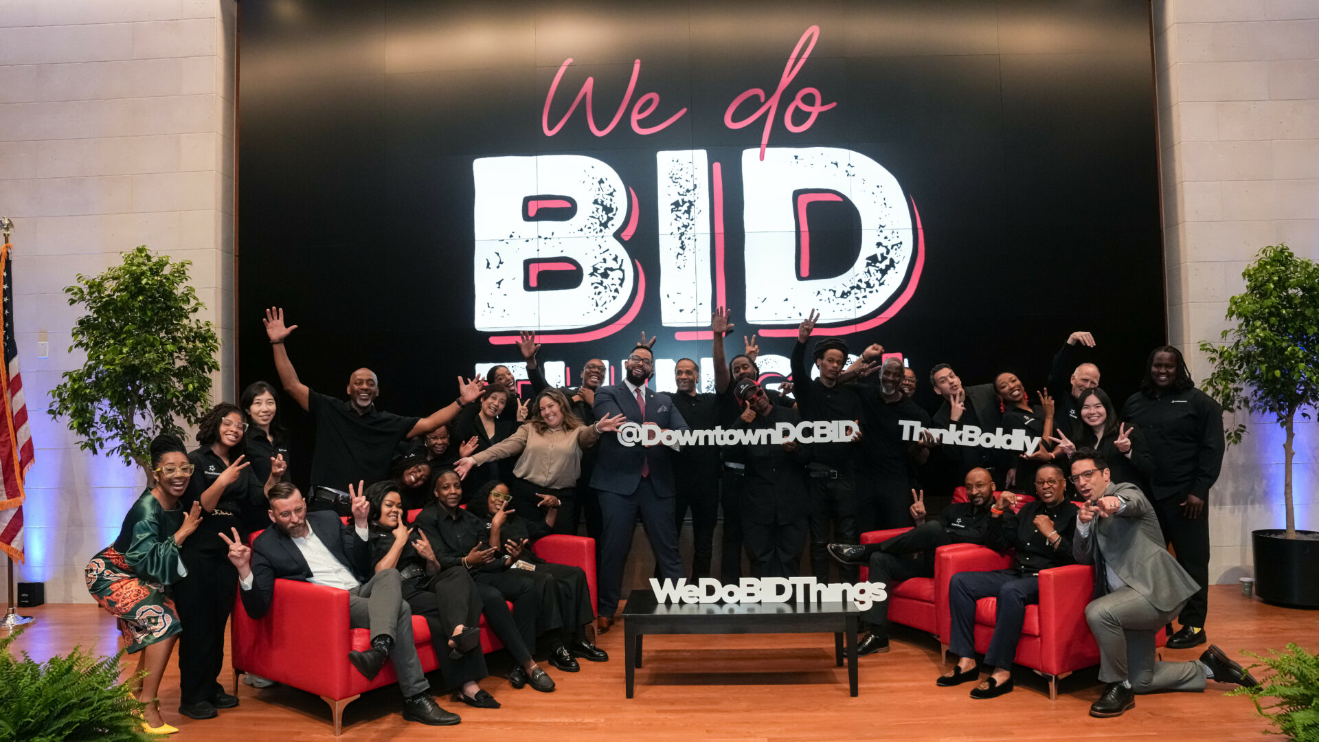 A large group of approximately 30 people, likely employees or members of a Business Improvement District (BID), posing for a lively group photo. Most people are wearing black and some are making celebratory gestures like peace signs or waving. They are clustered around two red armchairs and a small coffee table that reads "WeDoBIDThings". Behind them is a large black backdrop with the text "We do BID things" in white and pink script, along with social media handles like "@DowntownDCBID" and "#ThinkBoldly". An American flag and potted plants frame the scene.