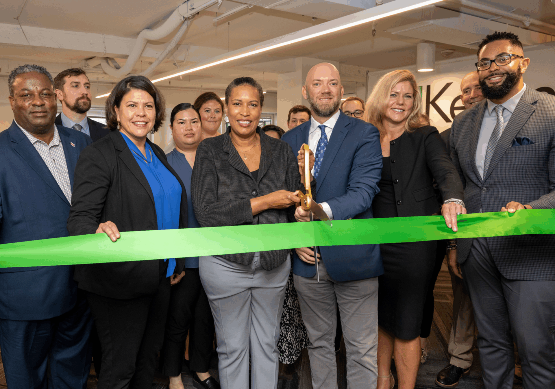 A diverse group of eight people, dressed in business attire, stands shoulder-to-shoulder, posing for a photo during a ribbon-cutting ceremony. A Black woman in a gray suit and a bald white man in a blue suit are positioned in the center, together holding a large pair of gold scissors to cut a thick, green ribbon that stretches across the group. Everyone is smiling, celebrating the event.