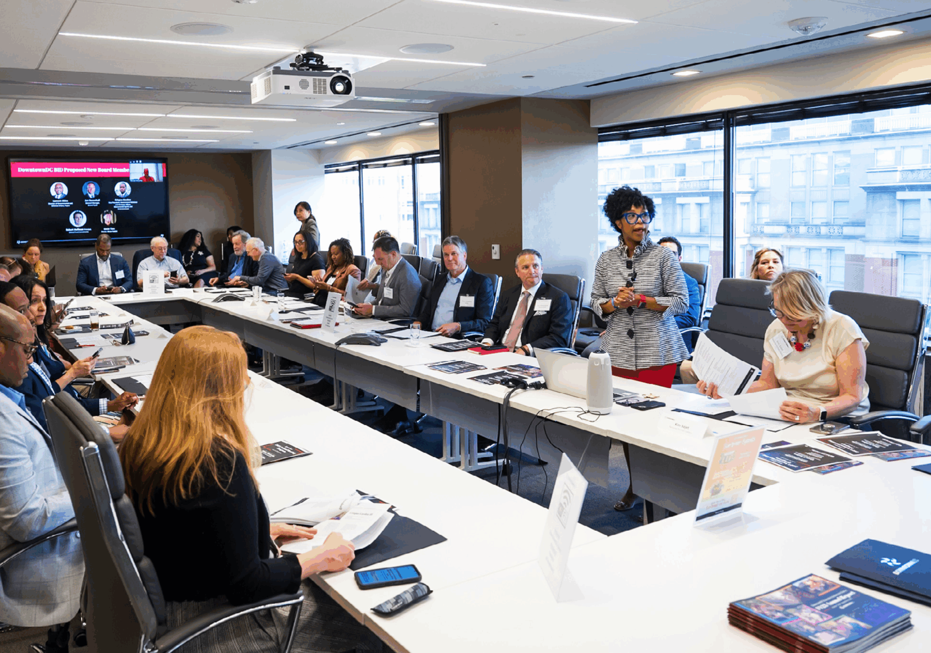 A professional photograph taken inside a modern, windowed conference room where a long U-shaped table is filled with people attending a meeting or presentation. A Black woman in a gray jacket and red skirt is standing and speaking at the corner of the table. Another woman in a white top is seated next to her, looking down at papers. A flat-screen monitor on the wall shows a digital presentation, and a projector hangs from the ceiling. The windows offer a view of city buildings outside.