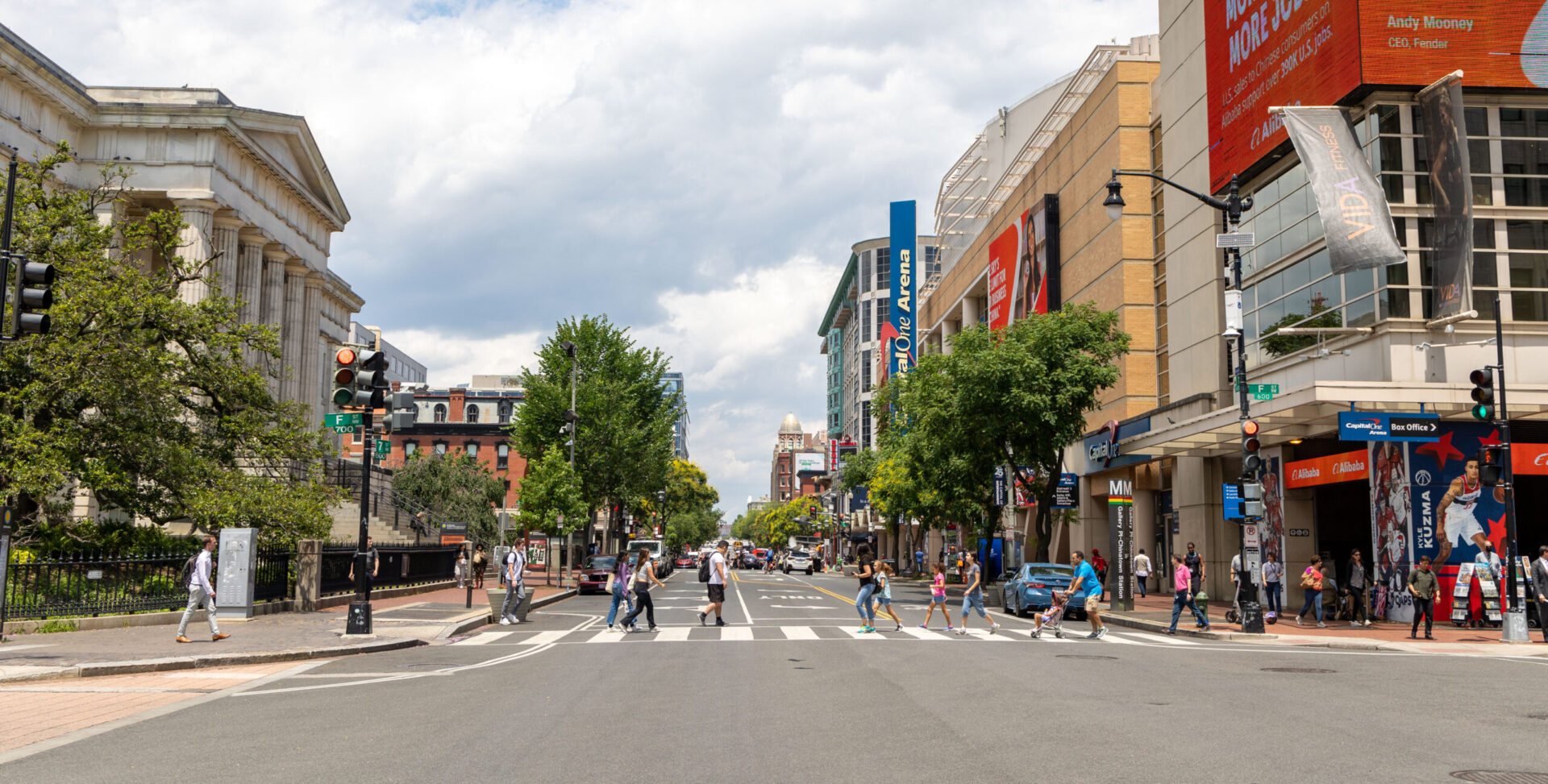 A bright, mid-day street scene of a busy downtown intersection with a crosswalk. Numerous pedestrians are crossing the street. On the left is a classical building with large columns. On the right are several modern commercial buildings with store signs and advertisements. Trees line the street, and a cloudy blue sky is visible overhead. The image has a soft-focus or blurred effect, especially on the periphery.