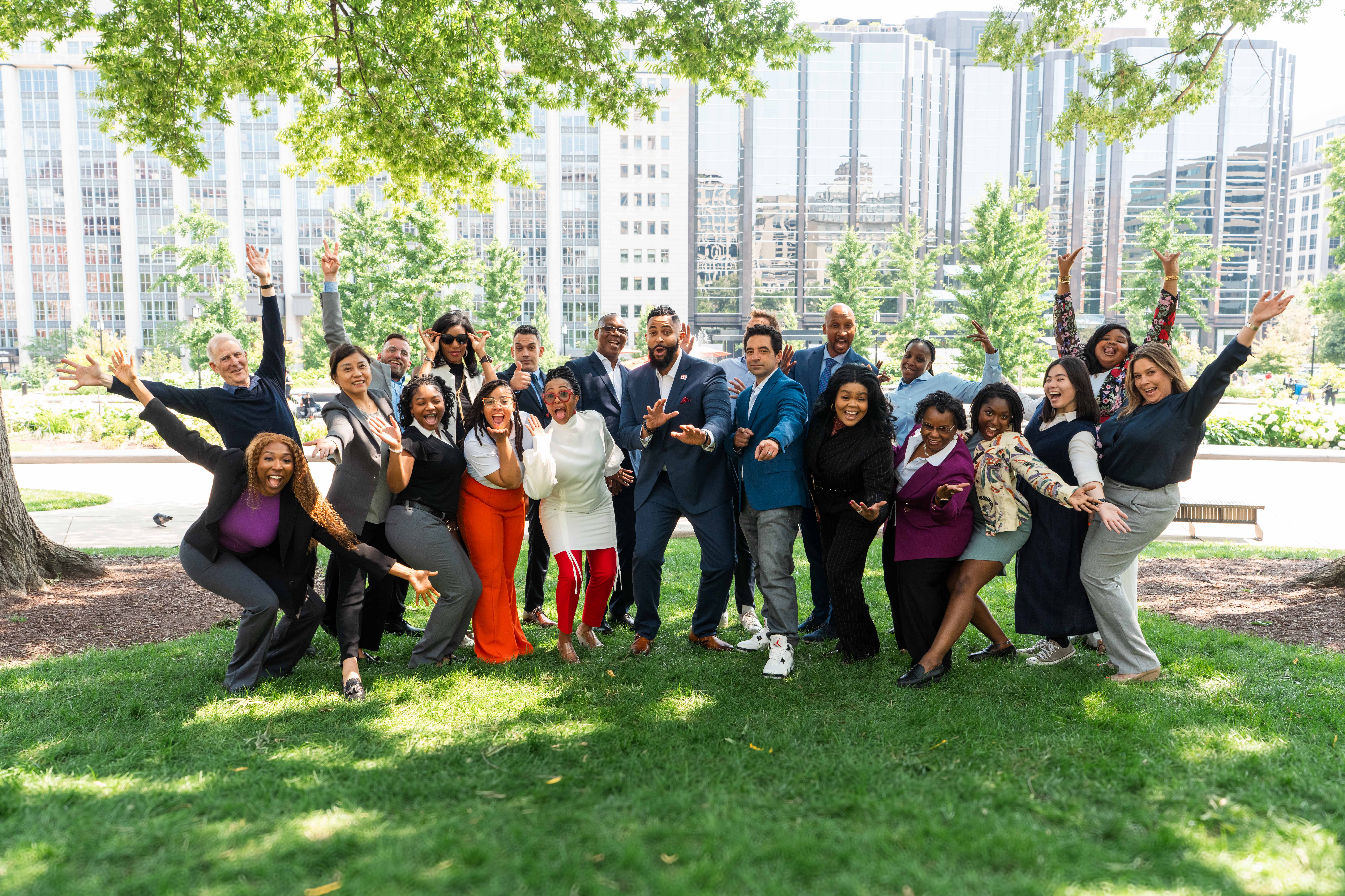 A diverse group of over twenty business professionals poses outdoors on a grassy lawn for a spirited group photo. Many people are smiling, raising their arms, and making dynamic poses. The group is in a city park setting, with trees providing shade in the foreground and tall, modern glass buildings visible in the sunny background.