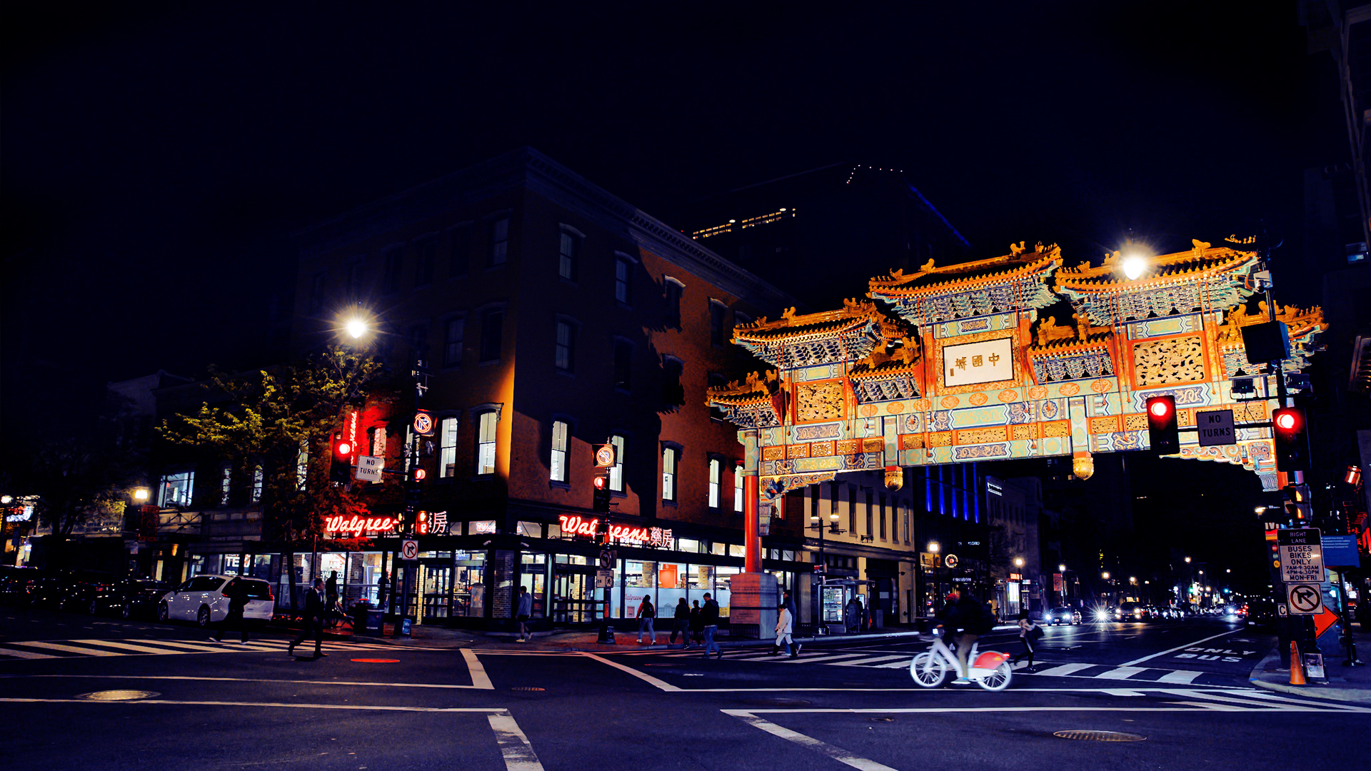 A nighttime street-level photograph of the Friendship Archway in Washington D.C.'s Chinatown. The large, colorful, and highly decorative arch is prominently featured in the center, brightly lit and spanning the street. Surrounding the arch are streetlights, cars, and the illuminated signs of nearby businesses and buildings, including one with the HOTEL sign visible in the background. Itineraries