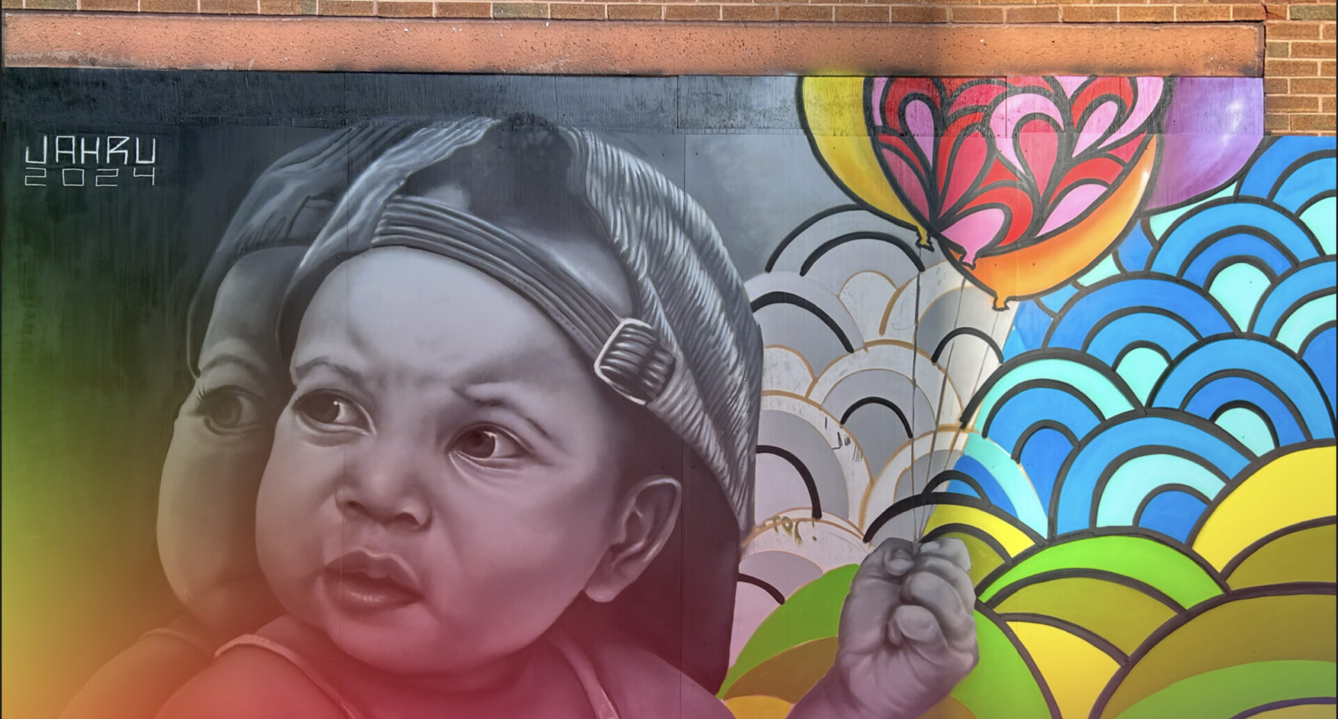 A close-up mural of a baby's face, primarily painted in black and white with a subtle shadow effect. The baby is wearing a baseball cap and has an expressive, somewhat scrunched or determined look. The right side of the mural features vibrant, colorful elements, including a brightly colored balloon and stylized waves or scallop shapes in blue, gray, yellow, and green. The artist's signature, "JAH.RU 2024," is visible in the upper left corner.