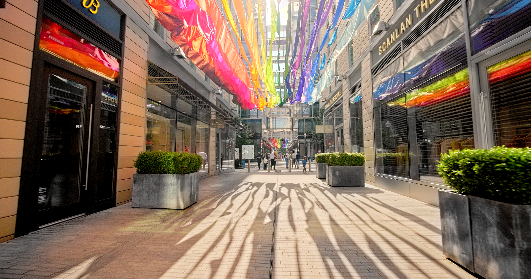 A photograph of an urban pedestrian walkway lined with glass-fronted shops. Colorful, translucent, rainbow-colored ribbons or fabric banners are suspended overhead, casting long, dramatic shadows onto the light-colored brick pavement below. Large concrete planters with trimmed green shrubs line the path. People are visible walking further down the brightly lit walkway. The sun shines brightly from above, highlighting the colors.