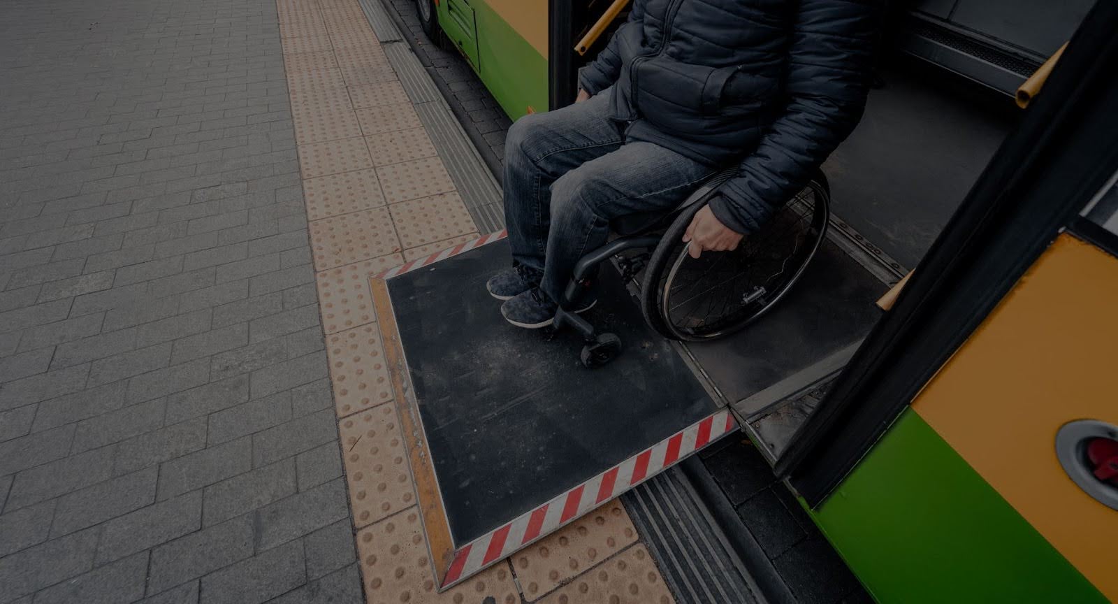 A man in a dark jacket is shown sitting in a wheelchair, moving onto or off of a public transit vehicle (likely a bus or streetcar) via a deployed accessibility ramp. The ramp bridges the gap between the vehicle and the tiled sidewalk, and the vehicle's door is open.