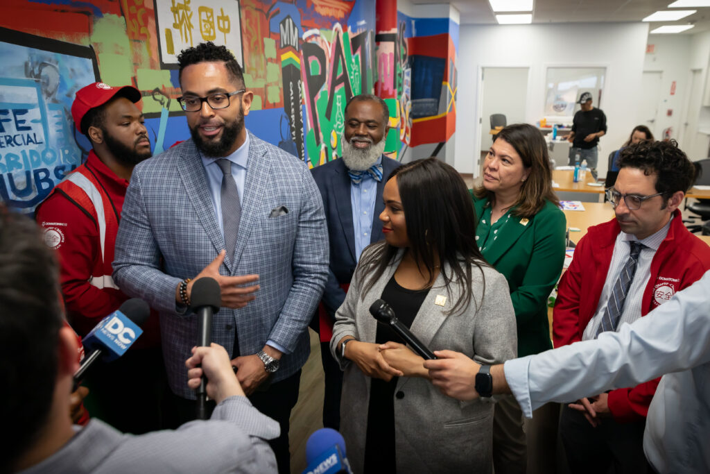 A group of people is gathered indoors in front of a colorful, graffiti-style mural. A well-dressed Black man in a light blue patterned suit jacket and glasses is speaking into several microphones held by reporters outside the frame. He is flanked by a man in a red uniform and a bearded man in a dark suit and a bow tie. A woman in a gray jacket and a woman in a green blazer stand to his right, smiling and listening. Another man in a red uniform and tie looks on from the far right.