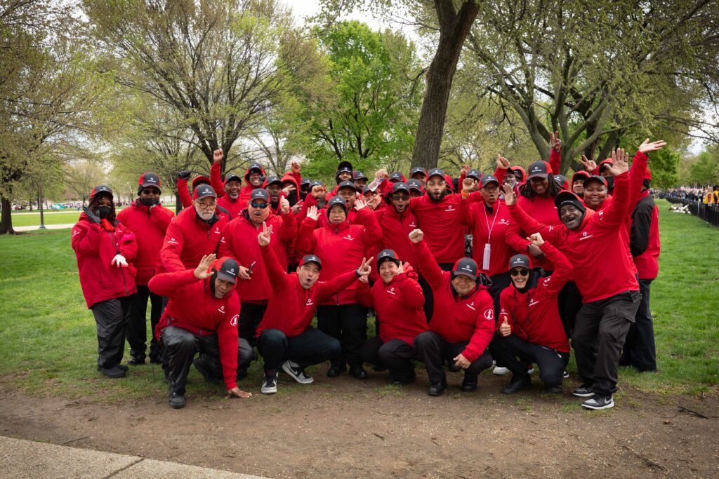 A large group of approximately 30 people, likely employees or volunteers, pose for a lively group photo outdoors in a park. All are wearing matching red jackets or sweatshirts and black caps. Many are smiling, cheering, or raising their hands in celebration. The group is standing and crouching in the center of the frame on a patch of dirt and grass, with trees and a large green lawn visible in the background.