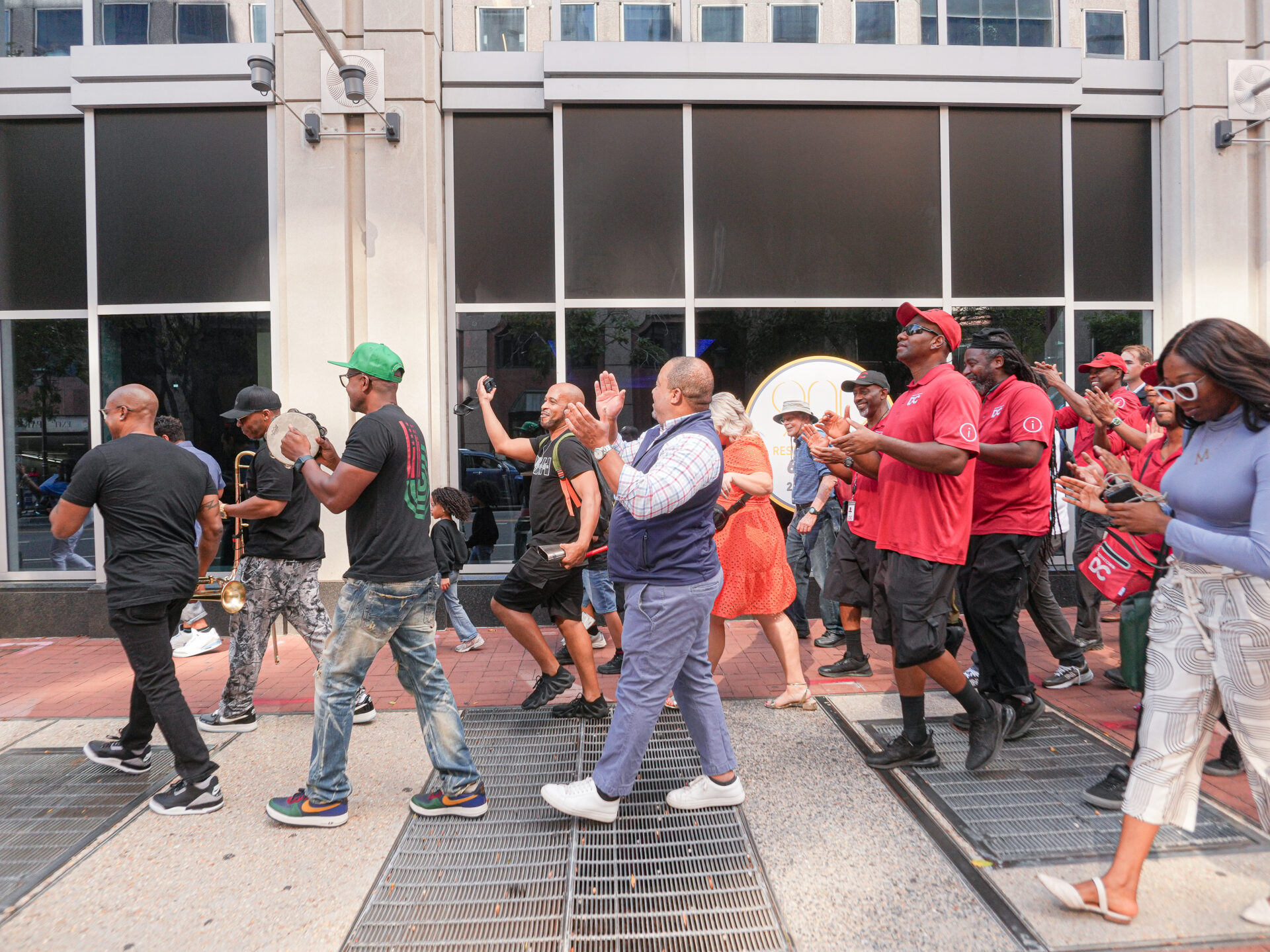 A diverse group of people is marching or walking in an organized group along a city street or sidewalk, possibly during a parade or demonstration. Many of the participants are clapping or cheering. Several men in the foreground are wearing matching red shirts and caps. The marchers are moving in front of a modern building with large, dark, reflective glass windows.