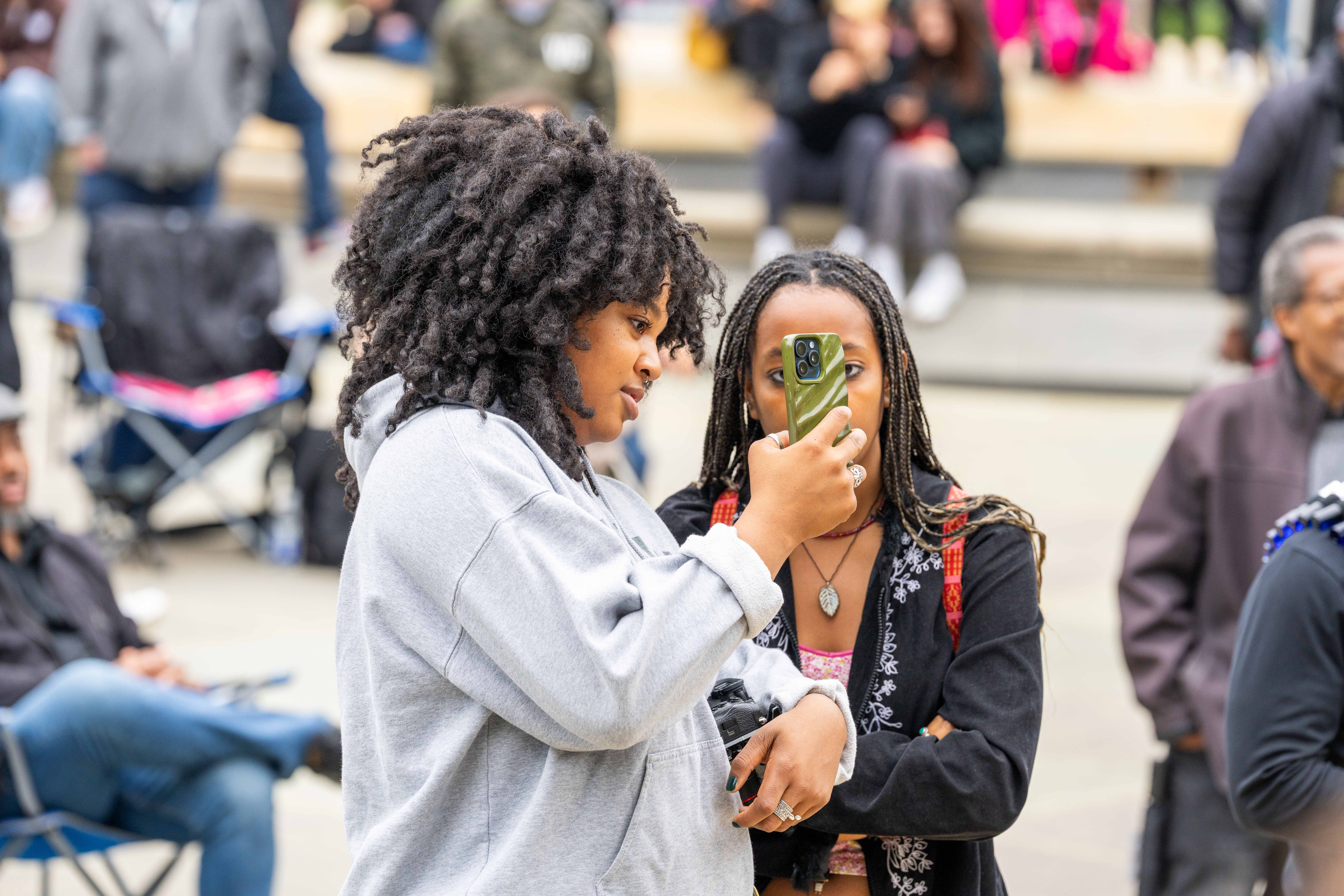 Two young women stand close together in an outdoor public space, likely at an event. The woman on the left, with large curly hair and a light gray sweatshirt, is holding a smartphone and showing the screen to the woman on the right. The woman on the right, with braided hair, is looking at the phone while holding her arm across her chest. Other people are visible, out of focus, in the background.
