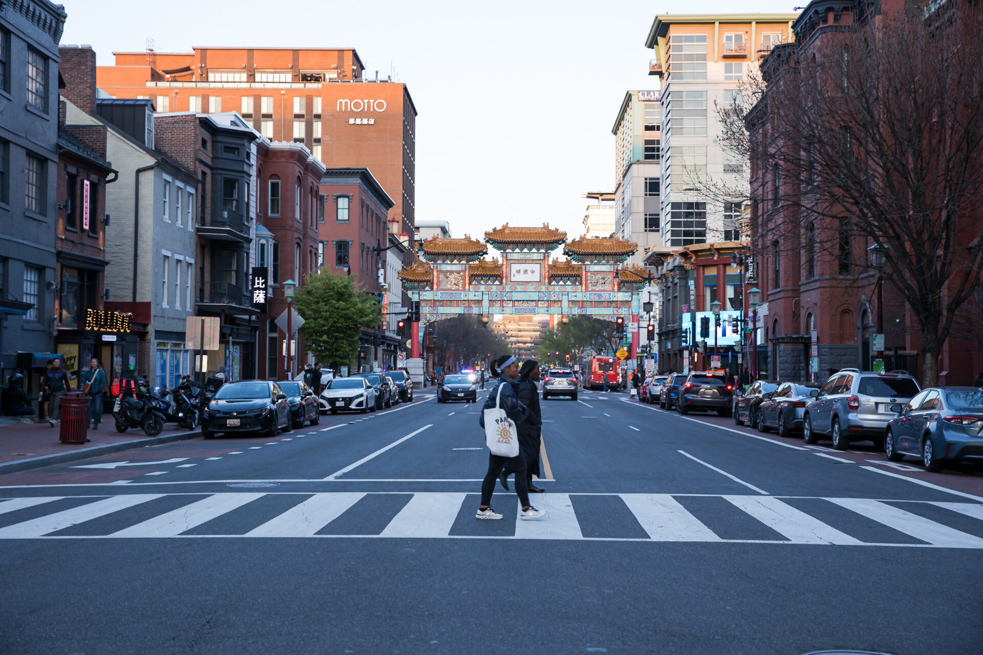 A daytime street-level photograph of a city street with the large, decorative Friendship Archway of Chinatown visible in the distance, spanning the road. The street is lined with parked cars and a mix of historic and modern buildings. Two pedestrians are walking across a white crosswalk in the foreground.