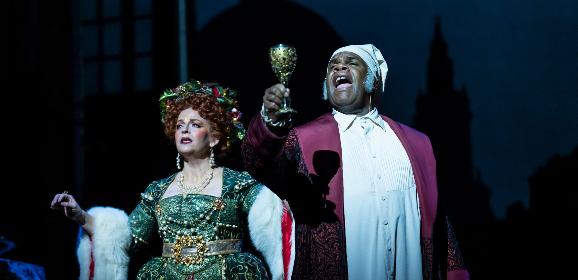 Two actors on a dark stage, wearing elaborate Victorian-era costumes. A Black man, likely playing the Ghost of Christmas Present, wears a red and white robe and holds up a gold chalice. A white woman in a green, bejeweled dress and a garland wreath stands next to him.