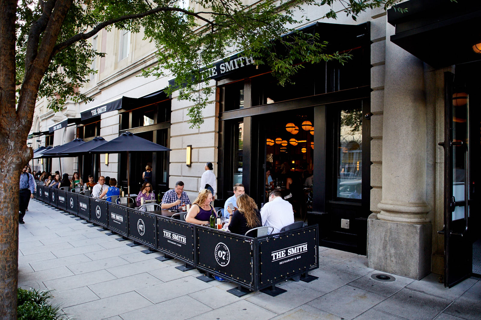 Outdoor tables line a busy street as groups of diners chat over meals in the open-air seating area.