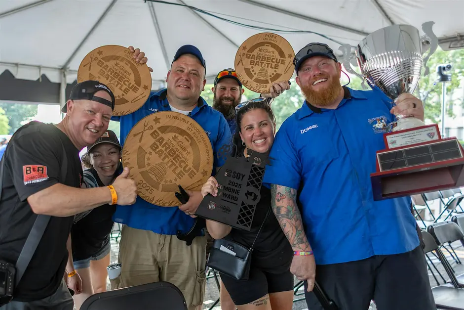 A group of smiling participants celebrates their victory at the Annual BBQ Battle, holding a large silver trophy and wooden award plaques while posing together on stage.