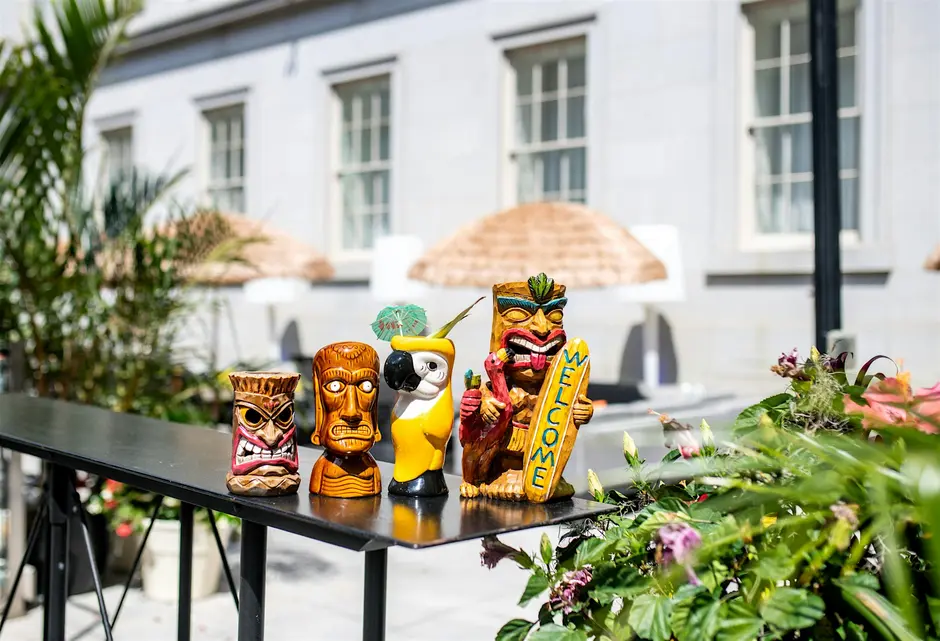 Four tropical, tiki-inspired cocktail glasses—three shaped like human faces and one like a yellow parrot—sit on a balcony overlooking an open-air courtyard. The Summer Affair: A Singles Night